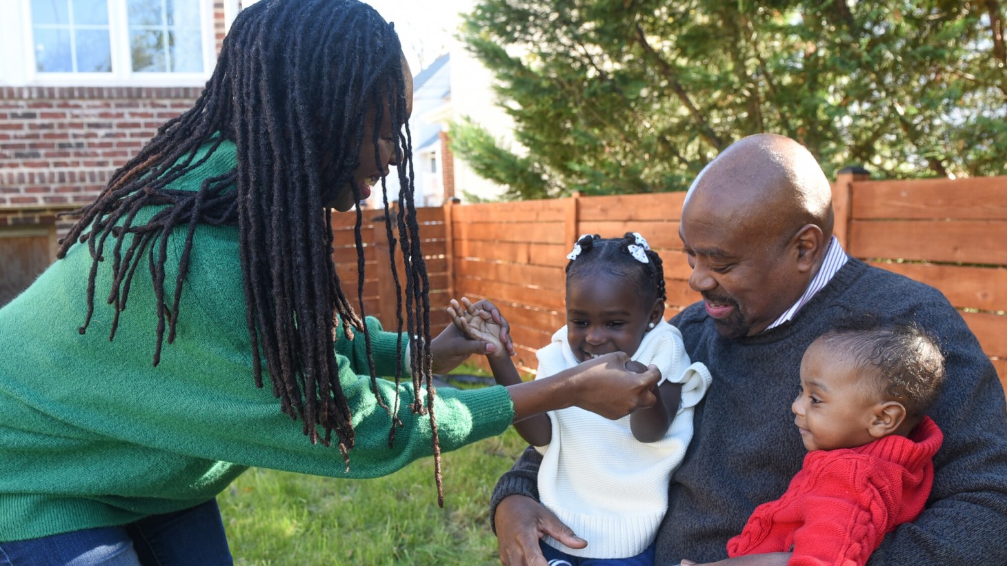 在一个阳光明媚的后院，一位黑人母亲牵着她的小女儿的手，她的小女儿和她的弟弟坐在爸爸的腿上。