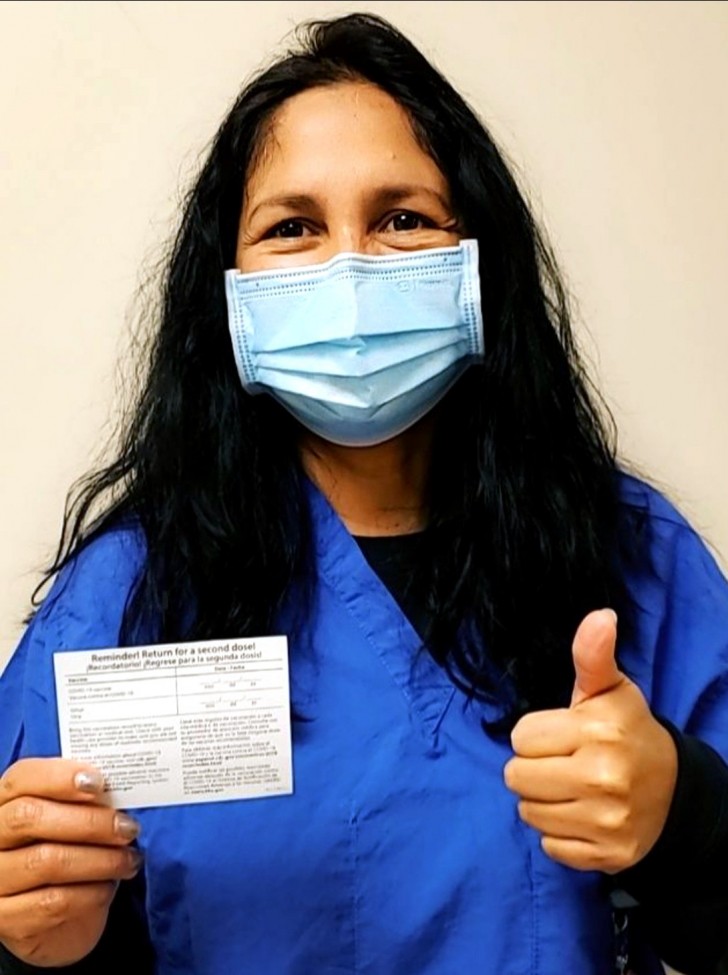 Woman with long black hair, wearing a mask, holding up vaccination card, giving thumbs up