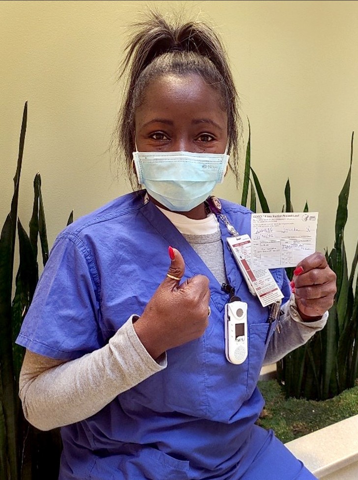 Black woman wearing blue scrubs, holding up her vaccination cards and giving the thumbs-up signal